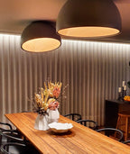 Two black dome Clérachic ceiling lights of different sizes glow over a long timber dining table with dried flowers and a pleated wall.
