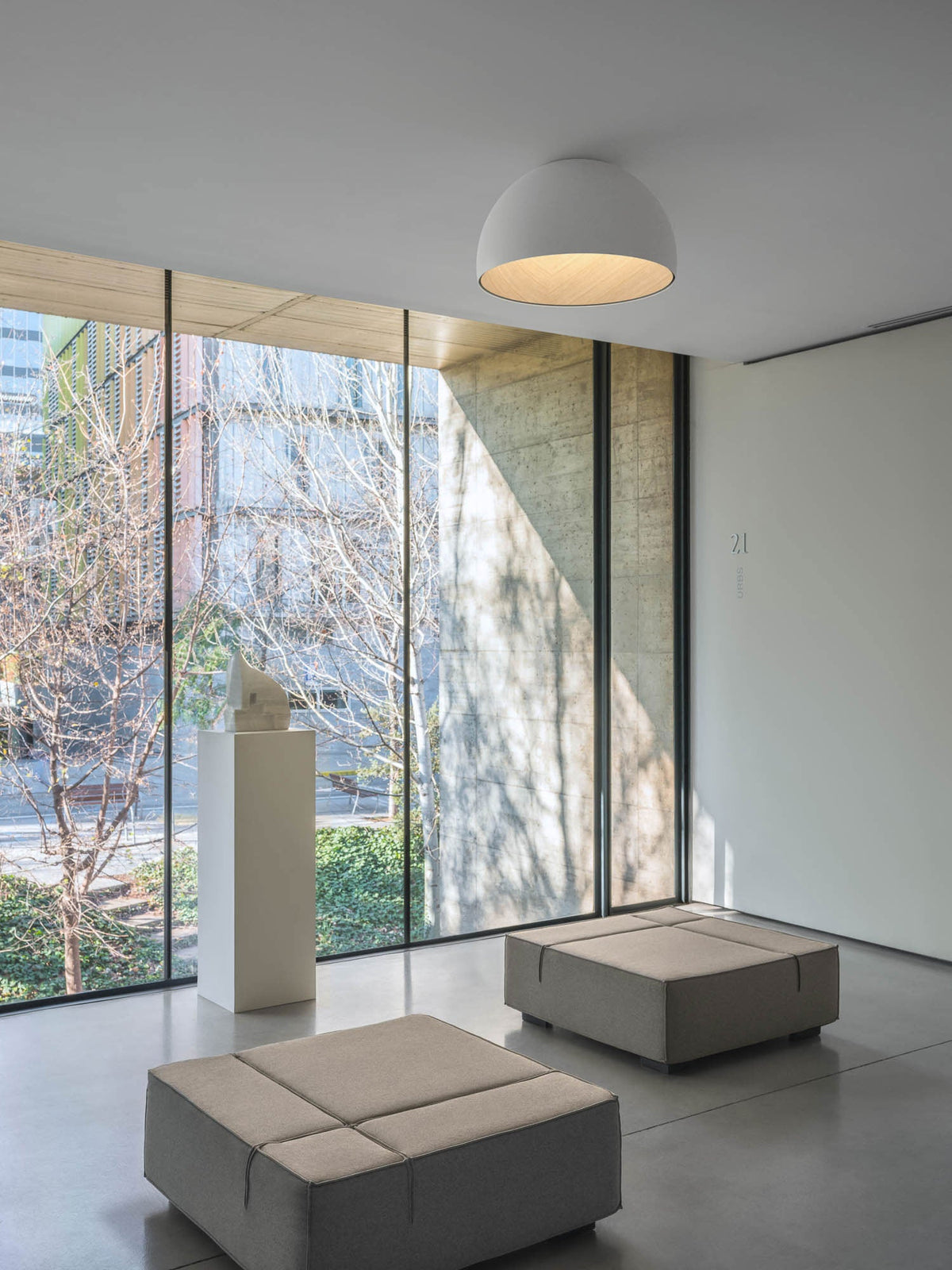 One white Clérachic dome ceiling light in a bare gallery room with floor-to-ceiling glazing, a plinth and two low grey ottomans.