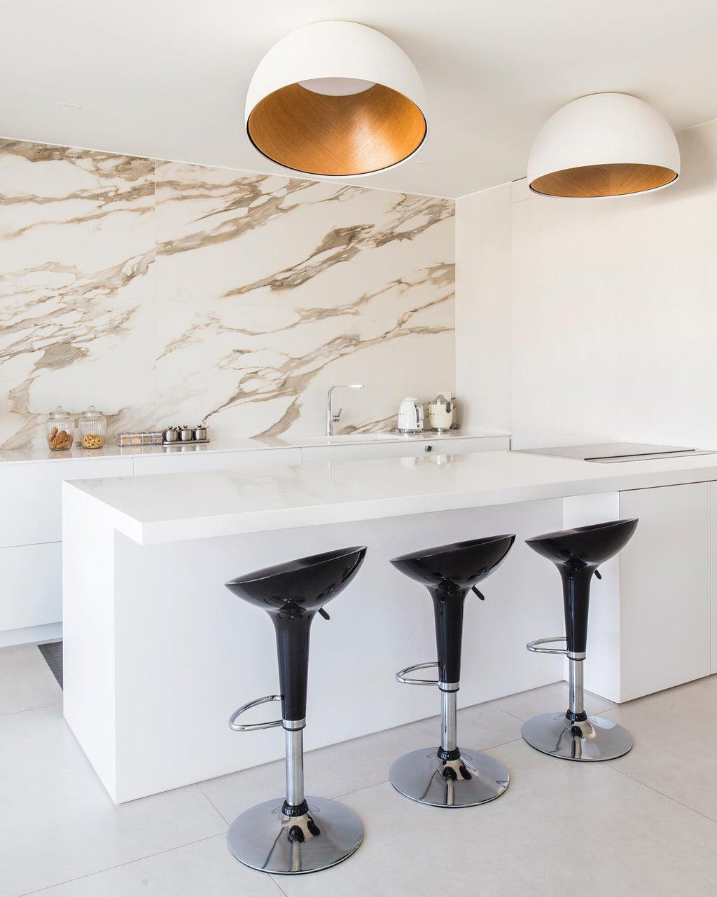 Two white domes with oak interiors, Clérachic ceiling lights, above a white kitchen island with black bar stools and a marble splashback.