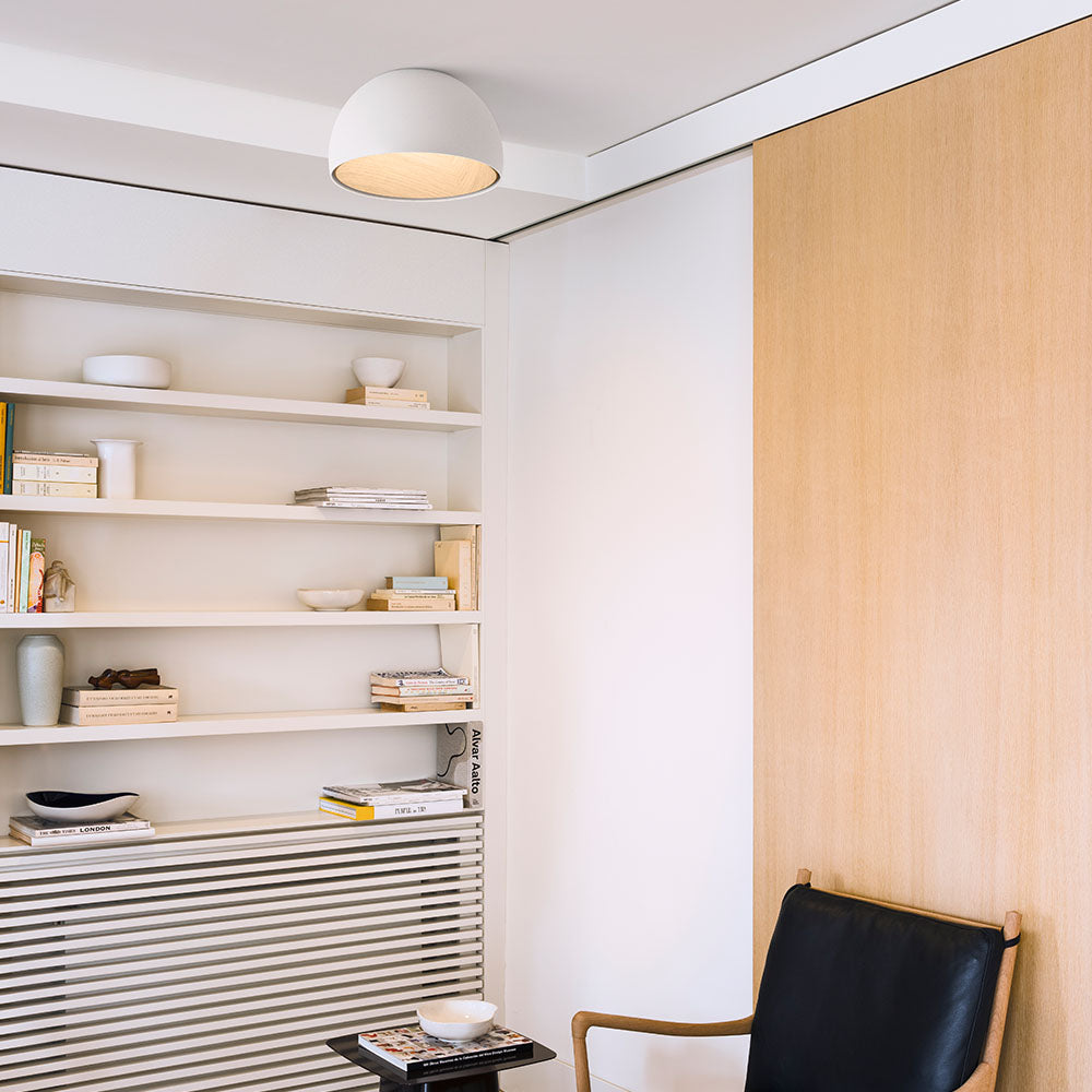 A white Clérachic dome ceiling light high in the corner of a room, with white shelves of books and a black leather chair below.