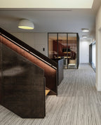 A small white Clérachic dome ceiling light lit warm above a corridor with a dark stair rail and a striped carpet.