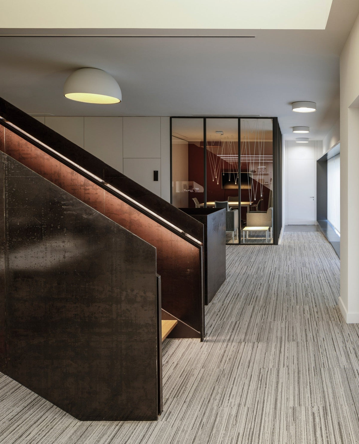 A small white Clérachic dome ceiling light lit warm above a corridor with a dark stair rail and a striped carpet.