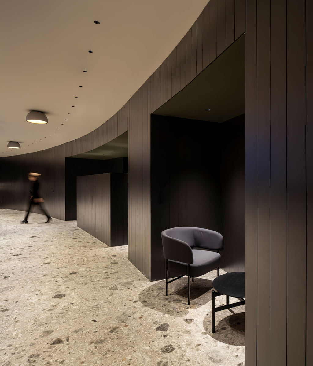 A small Clérachic dome ceiling light along a curved dark timber wall, beside a stone floor and a grey armchair.
