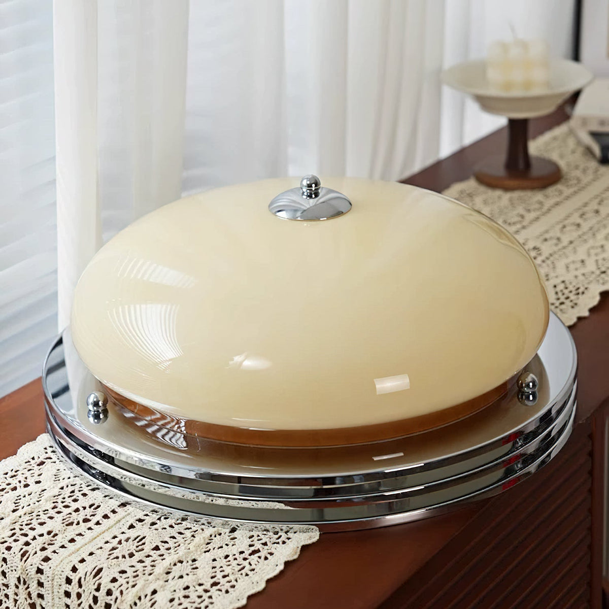 The Clérachic dome light resting on a lace runner on a wooden sideboard, showing its beige glass dome, chrome finial and stacked chrome rings.