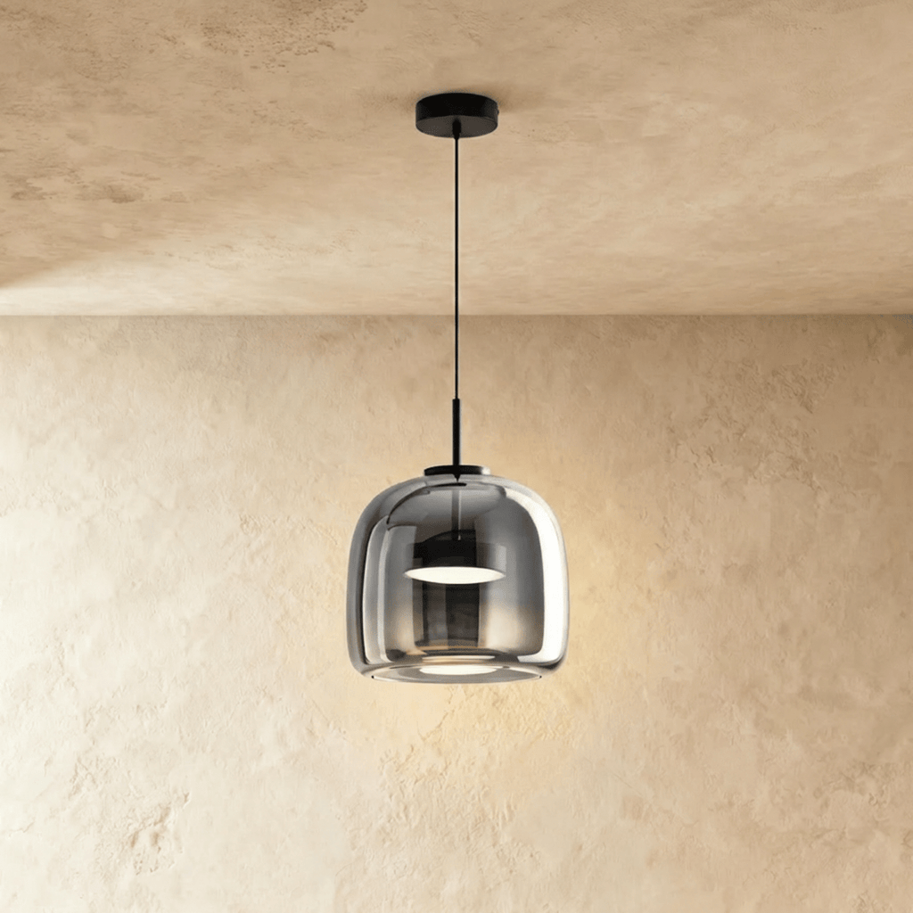 Clérachic smaller, rounder smoked glass pendant glowing softly on a black cord in front of a limewash wall.