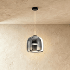 Clérachic smaller, rounder smoked glass pendant glowing softly on a black cord in front of a limewash wall.