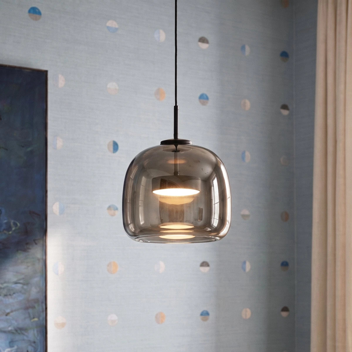 Clérachic close view of the rounded smoked glass pendant with a glowing disc diffuser, hung against pale blue wallpaper.