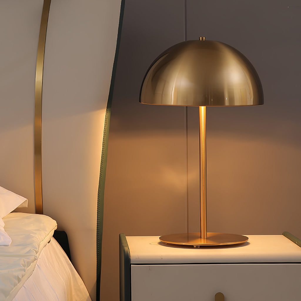 Brass-toned dome shade glowing warm on a bedside cabinet, a Clérachic table lamp beside a pillowed bed.