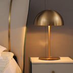 Brass-toned dome shade glowing warm on a bedside cabinet, a Clérachic table lamp beside a pillowed bed.