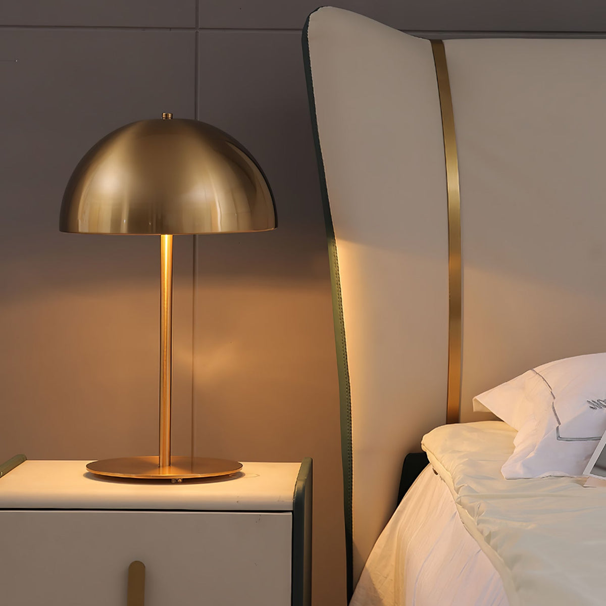 Closer view of a gold dome table lamp from Clérachic on a white bedside cabinet with a bed headboard behind.