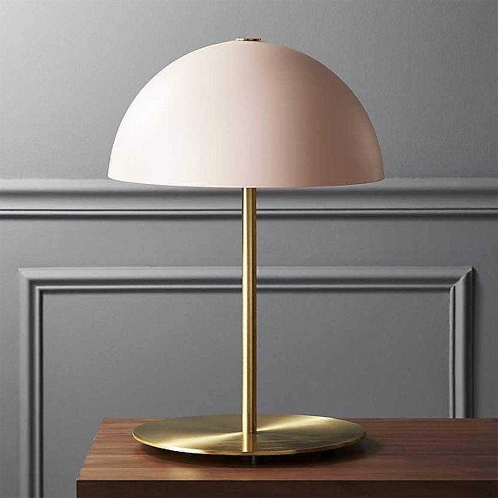Pink dome shade on a polished brass stem, a Clérachic table lamp on a dark wood table in front of grey panelling.
