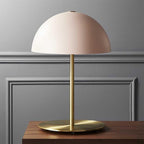Pink dome shade on a polished brass stem, a Clérachic table lamp on a dark wood table in front of grey panelling.