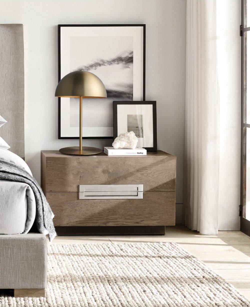 Gold dome table lamp from Clérachic on a grey wooden nightstand, beside framed prints and a pale bed.
