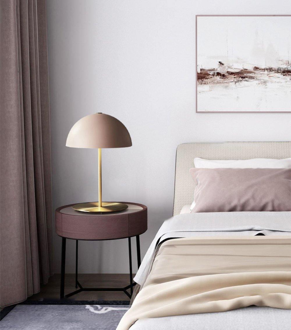 A Clérachic table lamp with a dusty pink dome shade on a round dark side table next to a bed with rose bedding.