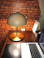 Clérachic dome table lamp with a brass base on a wooden desk, in front of a red brick wall and an open laptop.