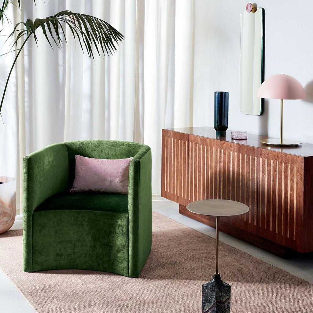 Pink dome lamp from Clérachic on a wooden sideboard, with a green velvet armchair and a tall plant in the room.