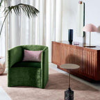 Pink dome lamp from Clérachic on a wooden sideboard, with a green velvet armchair and a tall plant in the room.
