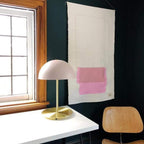 This Clérachic table lamp with a pale pink dome shade on a white desk by a paned window, with a wall hanging behind.