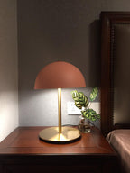 Terracotta-pink dome lamp from Clérachic on a dark wood bedside table, with a leafy plant and a wooden headboard.