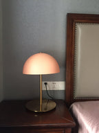 Lit dome table lamp from Clérachic with a peach-pink shade on a dark bedside table, with a wall socket visible.