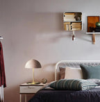 A Clérachic table lamp with a soft pink dome on a white nightstand, in a bedroom with an iron bedstead and shelf.