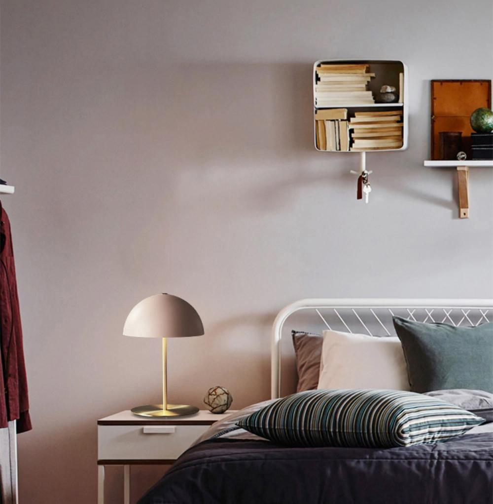 A Clérachic table lamp with a soft pink dome on a white nightstand, in a bedroom with an iron bedstead and shelf.