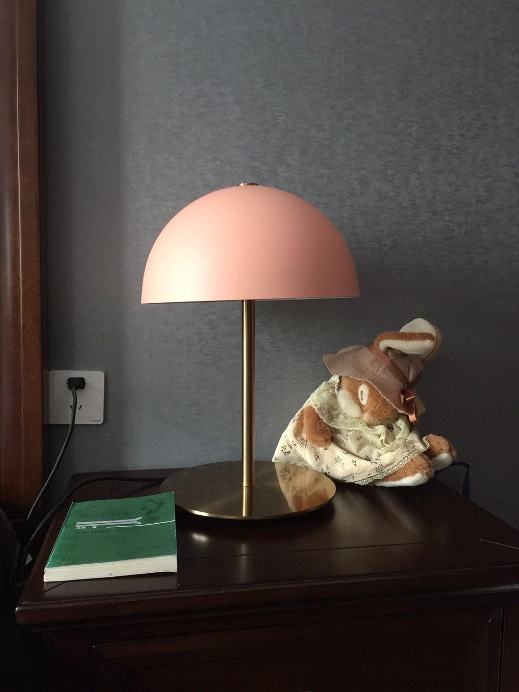 Coral-pink dome table lamp from Clérachic on a dark wooden table, beside a green book and a small soft toy.