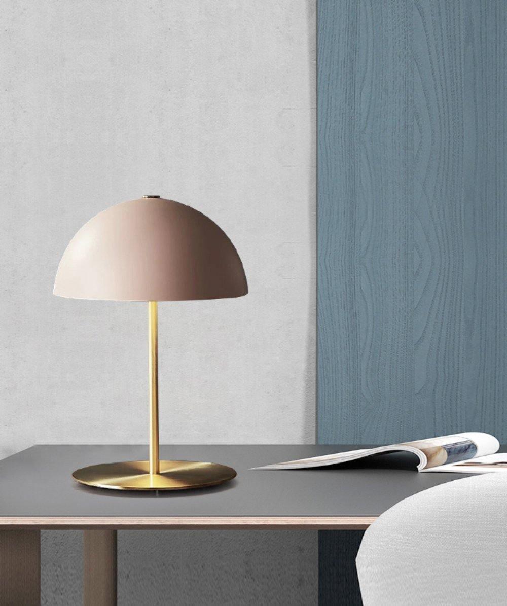 A Clérachic table lamp with a muted pink dome on a dark desk, with an open magazine and a blue-grey wall behind.