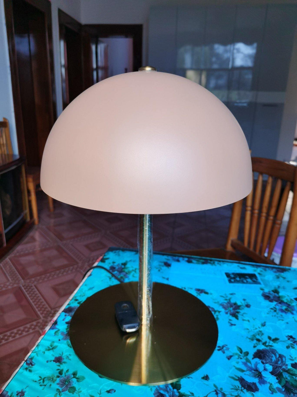 Customer-style view of a Clérachic table lamp with a pink dome shade, its brass base on a bright patterned tablecloth.