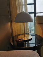 A white dome Clérachic table lamp lit on a round black side table, with a sofa and a curtained window behind.