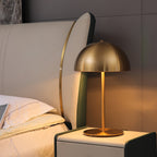 Lit brass dome table lamp from Clérachic on a bedside cabinet, with a cream headboard and white pillows behind.