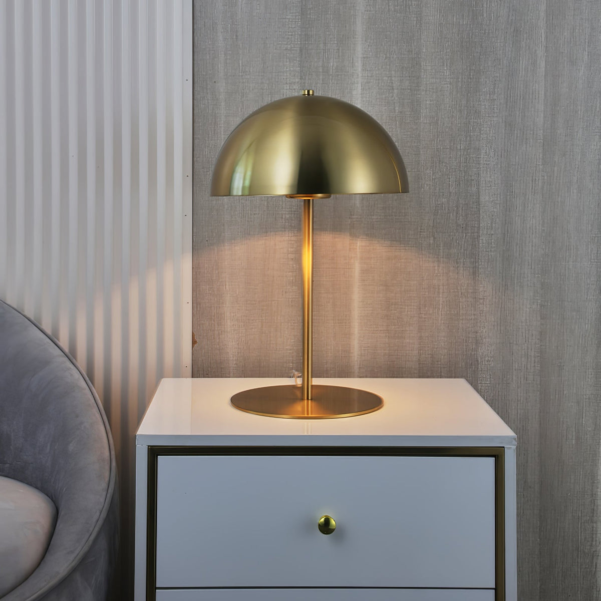 Gold dome table lamp from Clérachic on a white bedside chest, against a ribbed grey wall and a curved chair.