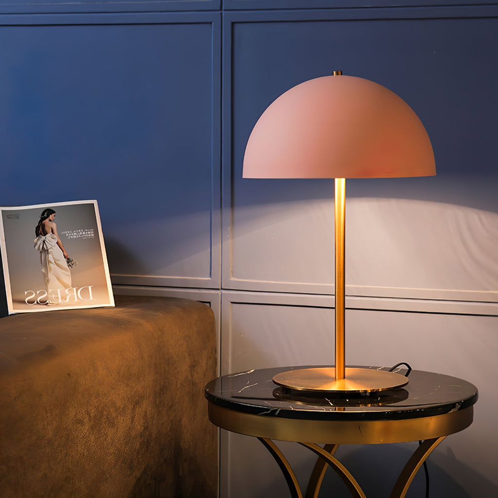 Pink dome lamp from Clérachic lit on a black marble table with a brass frame, with a magazine propped on blue panelling.