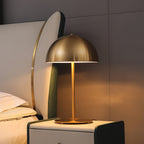 A Clérachic table lamp with a brass dome glowing warm on a white bedside cabinet, in front of a curved cream headboard.