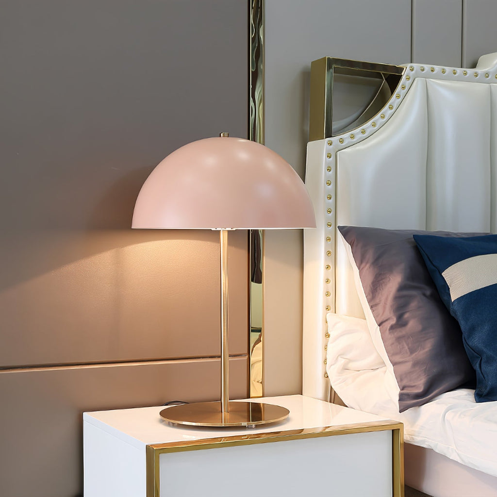 A Clérachic table lamp with a pink dome on a white nightstand, beside a bed with a studded cream headboard.