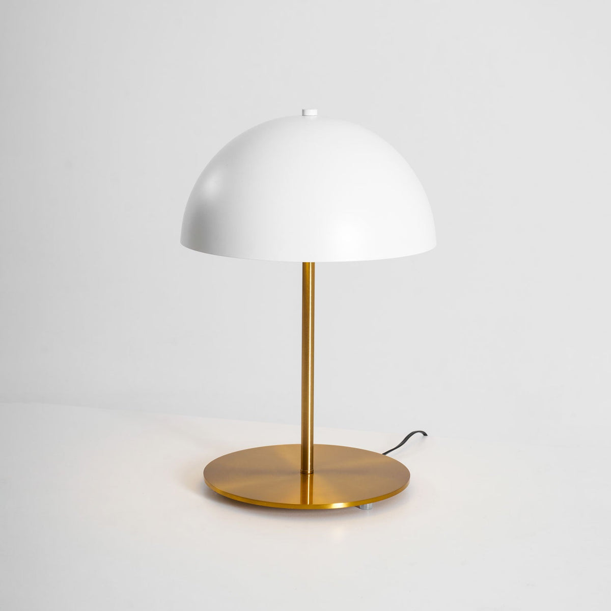 White dome table lamp from Clérachic with a brass base and a short black cord, on a pale grey backdrop.