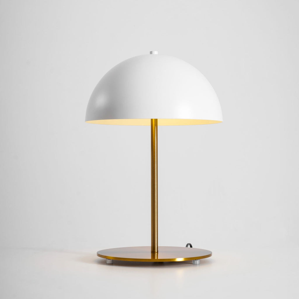 Lit white dome table lamp from Clérachic with a gold stem, its warm glow reflected in the base on a grey backdrop.