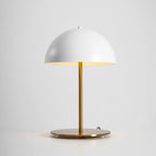 Lit white dome table lamp from Clérachic with a gold stem, its warm glow reflected in the base on a grey backdrop.