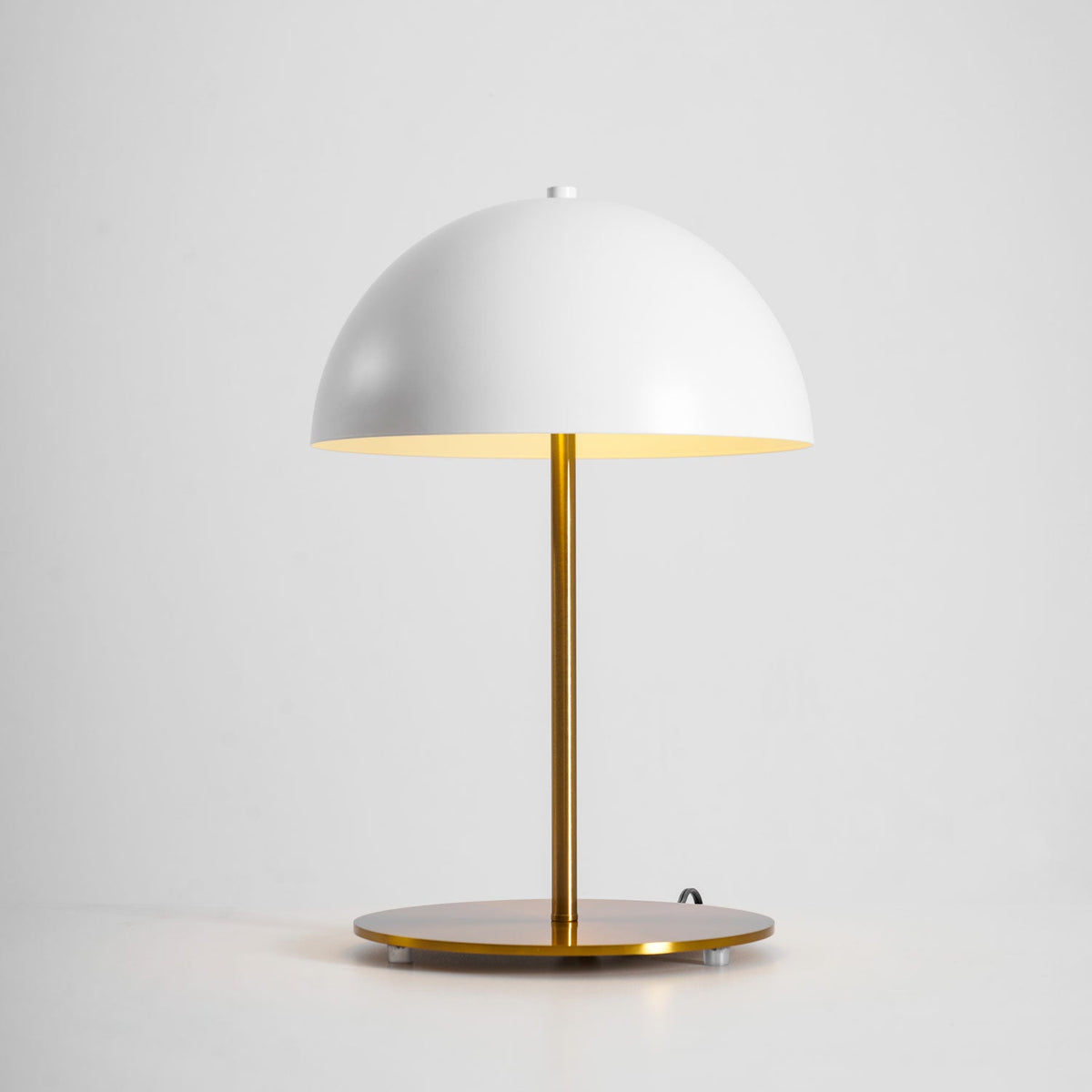 Lit white dome table lamp from Clérachic with a gold stem, its warm glow reflected in the base on a grey backdrop.
