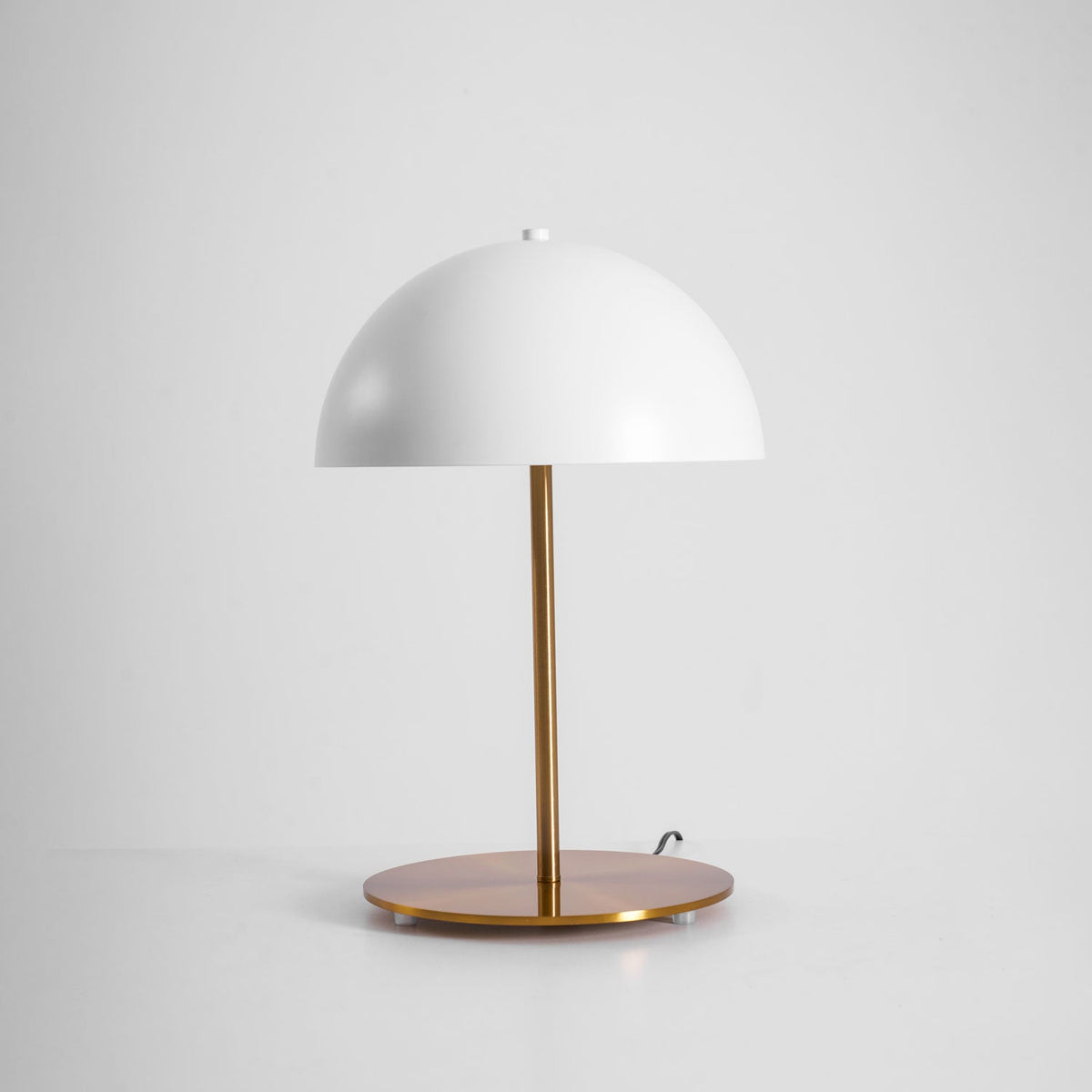 White dome table lamp from Clérachic seen from slightly above, with its round brass base and cord on a light backdrop.