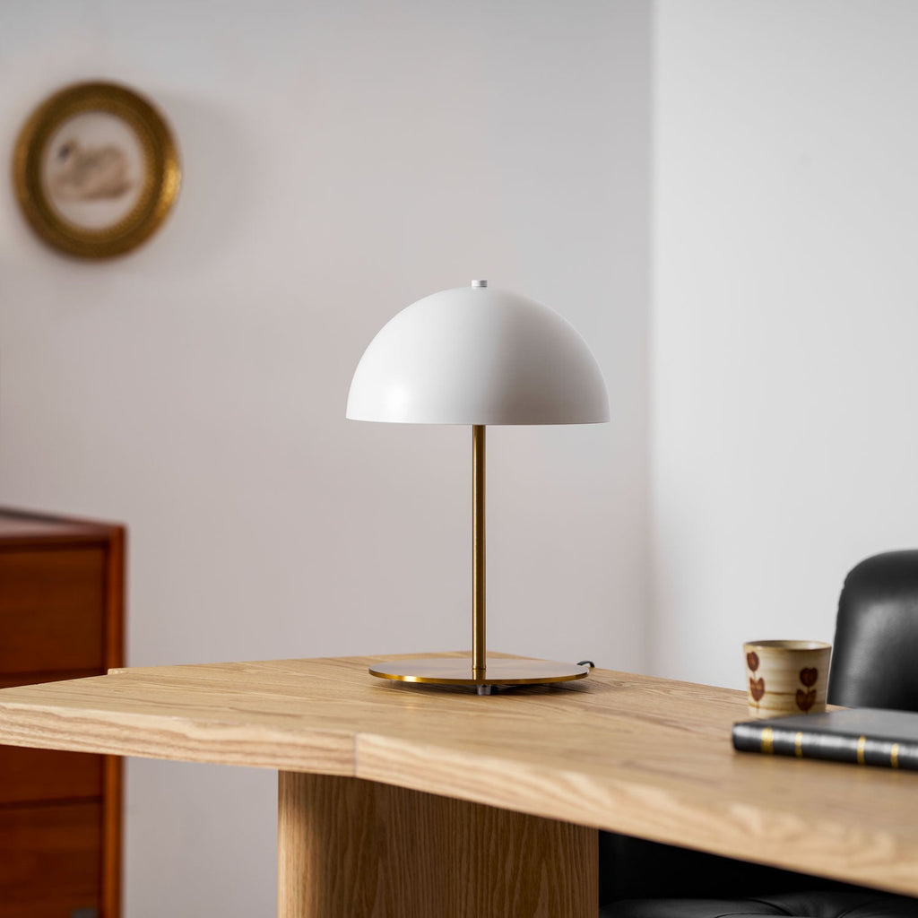 Small white dome table lamp from Clérachic on a pale oak desk, with a gilt wall plate and a patterned cup nearby.