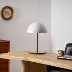 Small white dome table lamp from Clérachic on a pale oak desk, with a gilt wall plate and a patterned cup nearby.