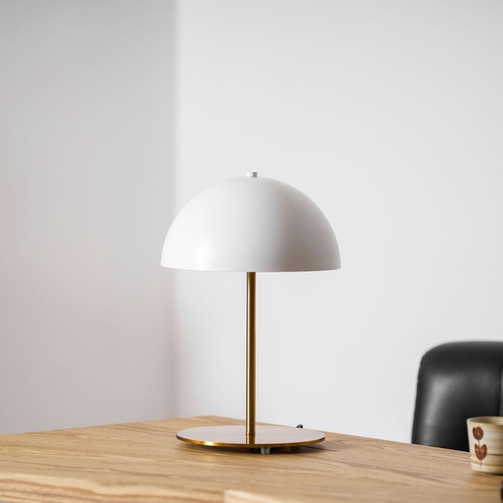 Closer view of a white Clérachic table lamp with a gold stem on a pale oak desk, beside a black leather chair.