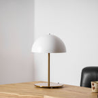 Closer view of a white Clérachic table lamp with a gold stem on a pale oak desk, beside a black leather chair.