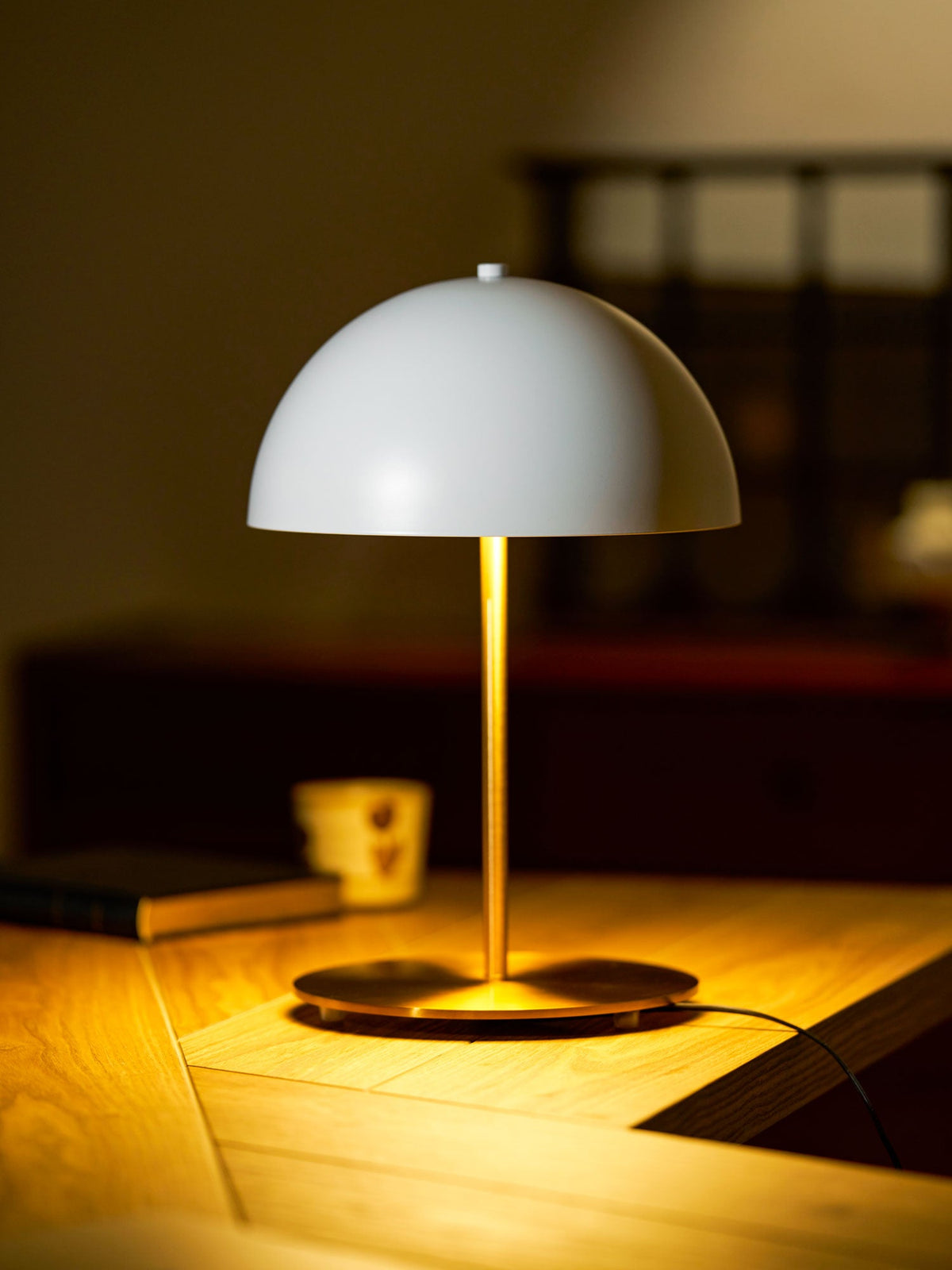 Warmly lit white dome table lamp from Clérachic casting a golden pool of light across a wooden desk in a dim room.