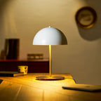 A Clérachic white dome lamp glowing gold on a wooden desk, with books, a cup and a round wall plate in the background.