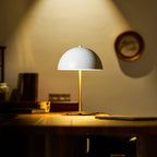 White dome table lamp from Clérachic lit on a dim desk, light spilling up the wall behind and a closed laptop beside it.