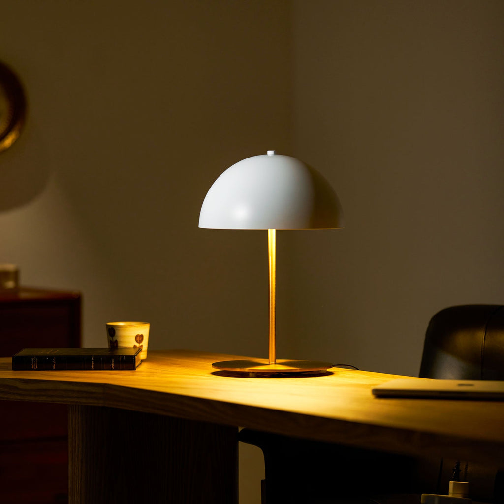 Evening scene with a white dome Clérachic table lamp lit on a wooden desk, a patterned cup and books beside it.