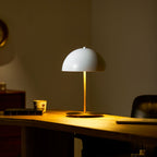 Evening scene with a white dome Clérachic table lamp lit on a wooden desk, a patterned cup and books beside it.