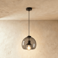 Clérachic single pendant with a smoked glass shade, a black cord and ceiling rose, its filament bulb lit against limewash.